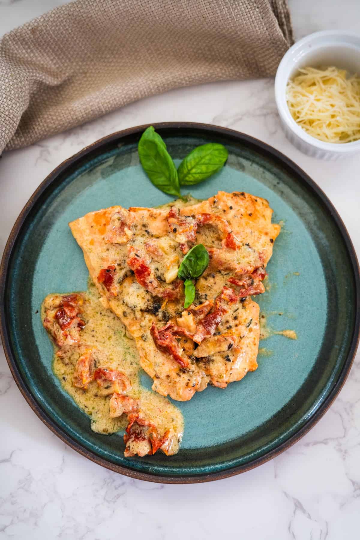 Grilled salmon topped with a creamy sauce and sun-dried tomatoes on a blue plate, garnished with basil leaves. A small bowl of grated cheese is in the background.