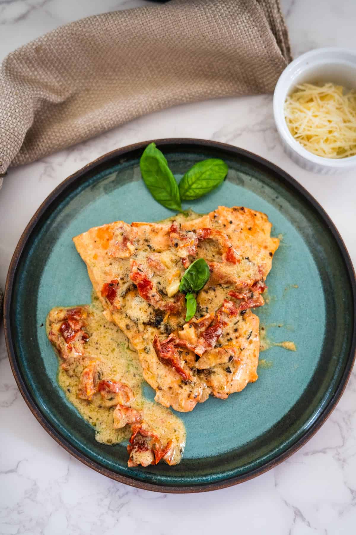 A plate of creamy sun-dried tomato chicken garnished with basil leaves, accompanied by a small bowl of grated cheese.