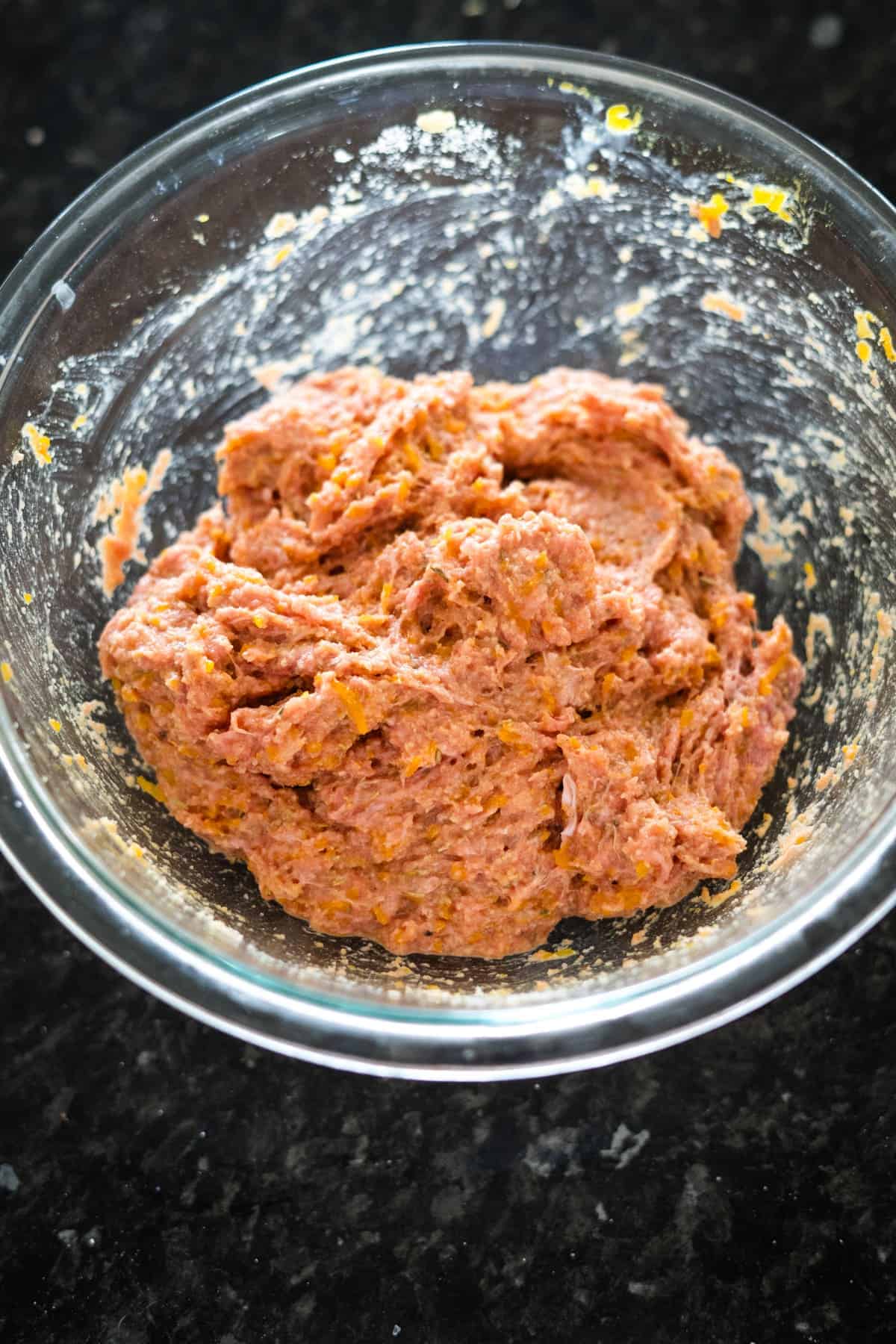 A glass bowl containing a mixture of minced meat and spices on a dark countertop.