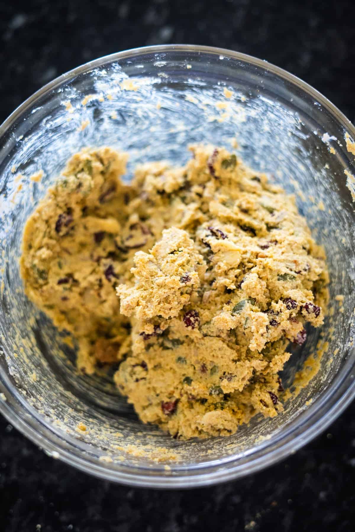 A glass bowl containing raw cookie dough mixed with chocolate chips on a dark countertop.