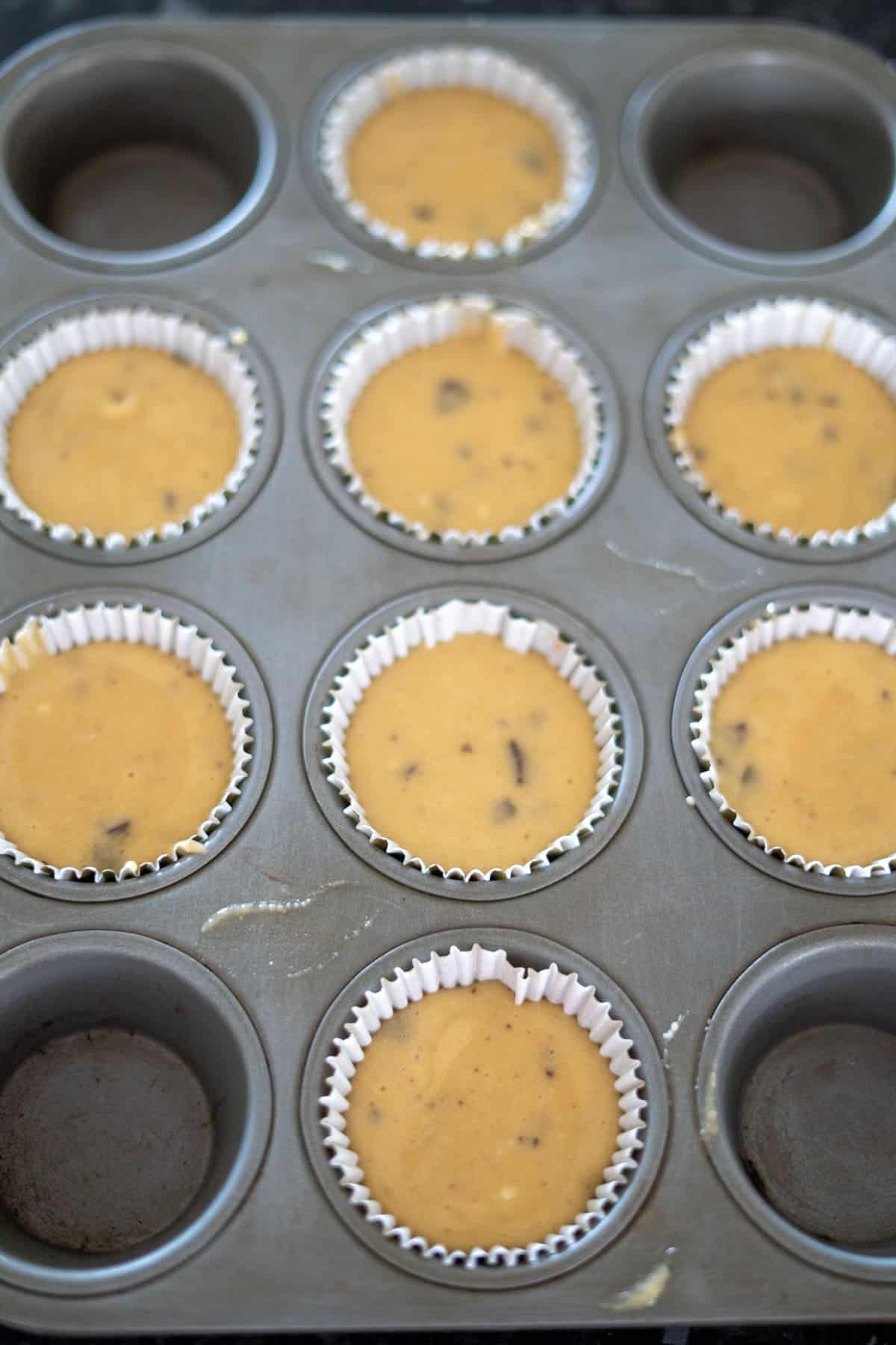 A muffin tin filled with nine paper liners, each containing chocolate chip batter, with three empty slots.