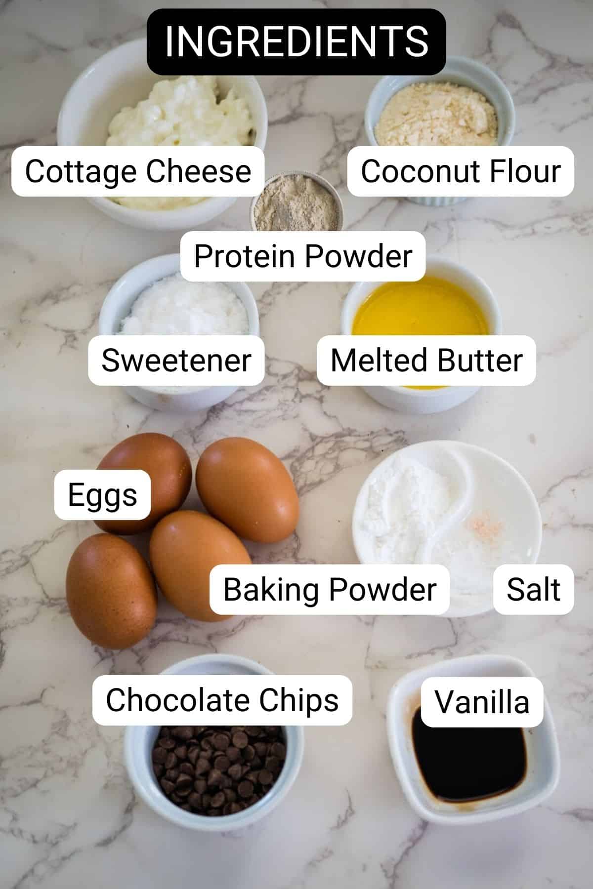 Labeled ingredients on a marble surface: cottage cheese, coconut flour, protein powder, sweetener, melted butter, eggs, baking powder, salt, chocolate chips, vanilla.