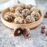 A round wooden plate with chocolate hazelnut truffles, some halved, and a few whole hazelnuts scattered on a gray surface.