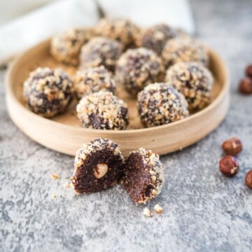 A round wooden plate with chocolate hazelnut truffles, some halved, and a few whole hazelnuts scattered on a gray surface.