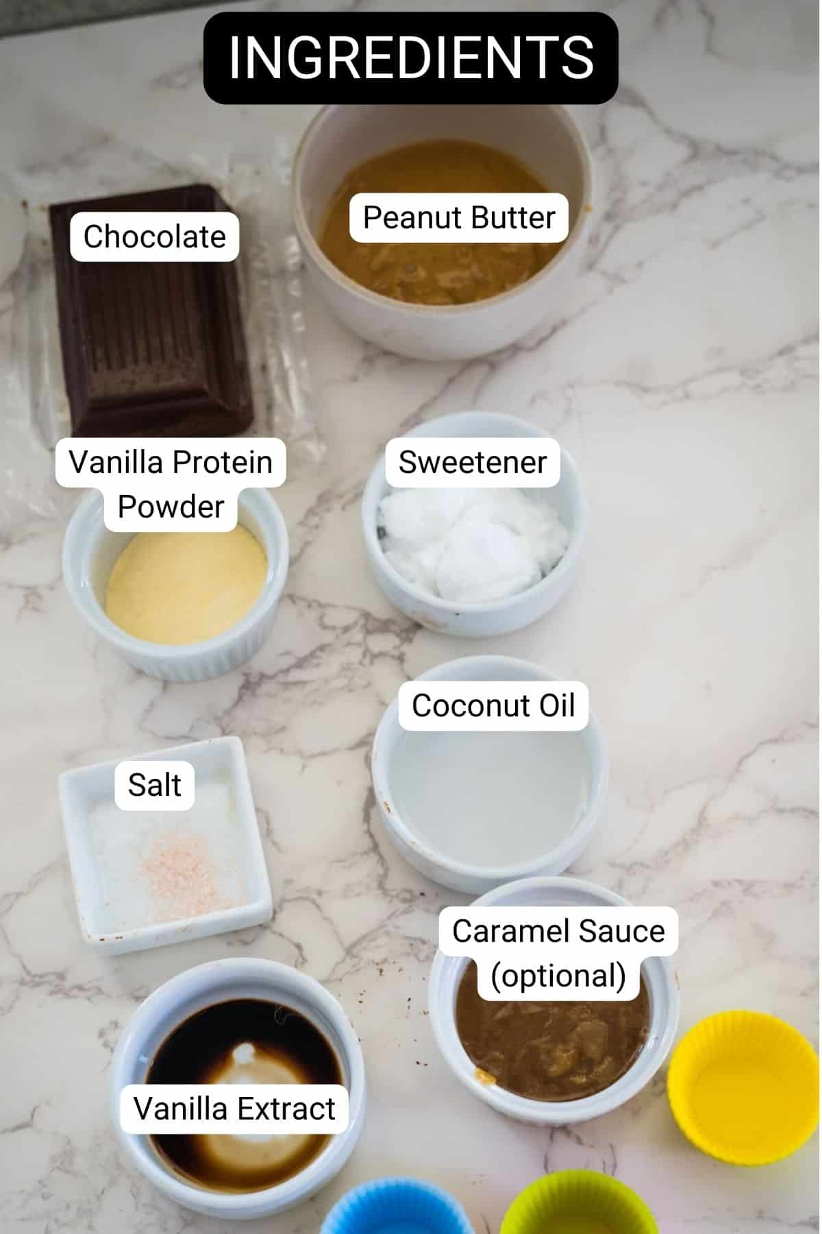 Ingredients including chocolate, peanut butter, vanilla protein powder, sweetener, coconut oil, salt, vanilla extract, and optional caramel sauce arranged on a marble surface.