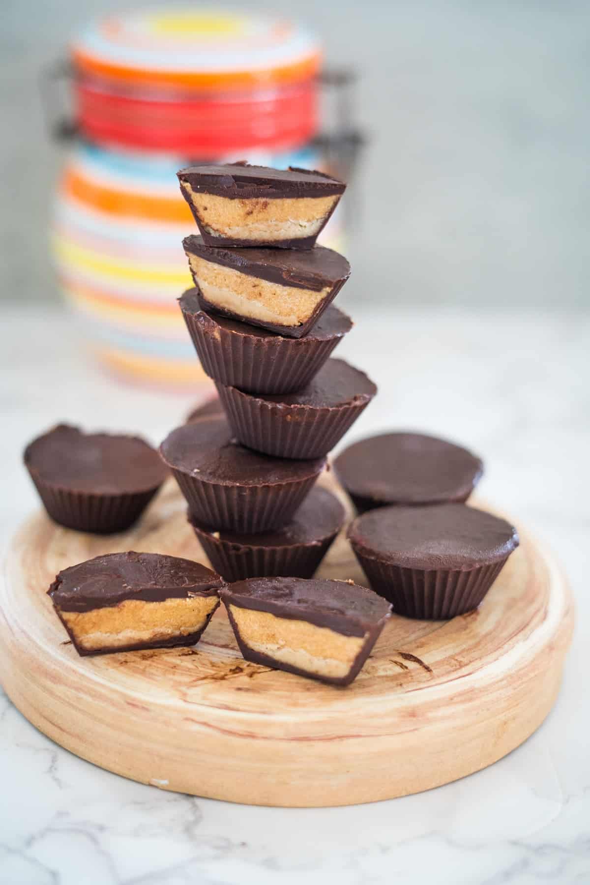 A stack of high protein chocolate peanut butter cups sits on a wooden board, with a colorful jar in the background.