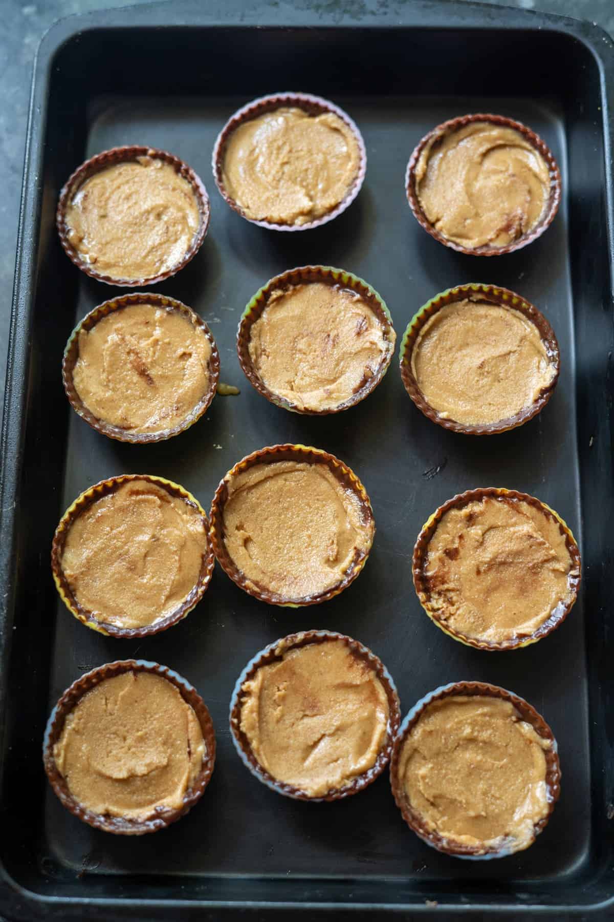 A baking tray with twelve unbaked muffin molds filled with creamy batter, ready to become delicious high-protein peanut butter cups.