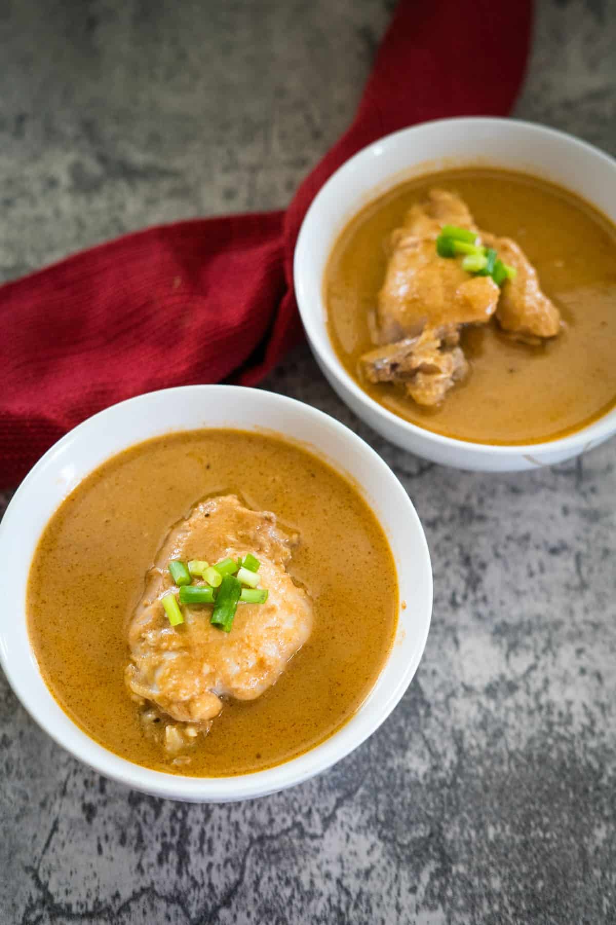 Two white bowls of soup, each containing a chicken piece and garnished with chopped green onions, are placed on a textured surface with a red cloth near them.