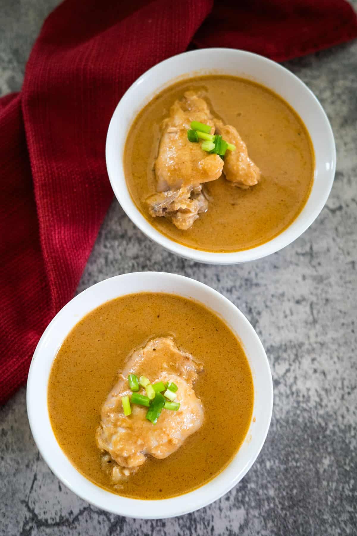 Two white bowls of curry with chicken pieces, garnished with chopped green onions, on a textured gray surface. Red cloth in the background.