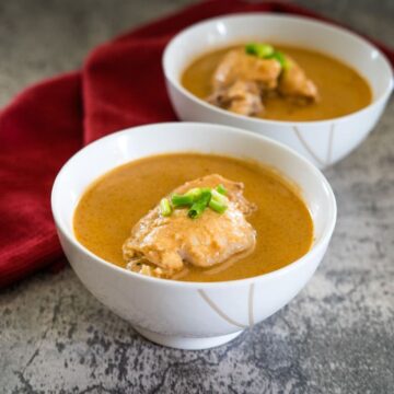 Two bowls of soup with chicken pieces, garnished with green onions, placed next to a red cloth on a textured surface.