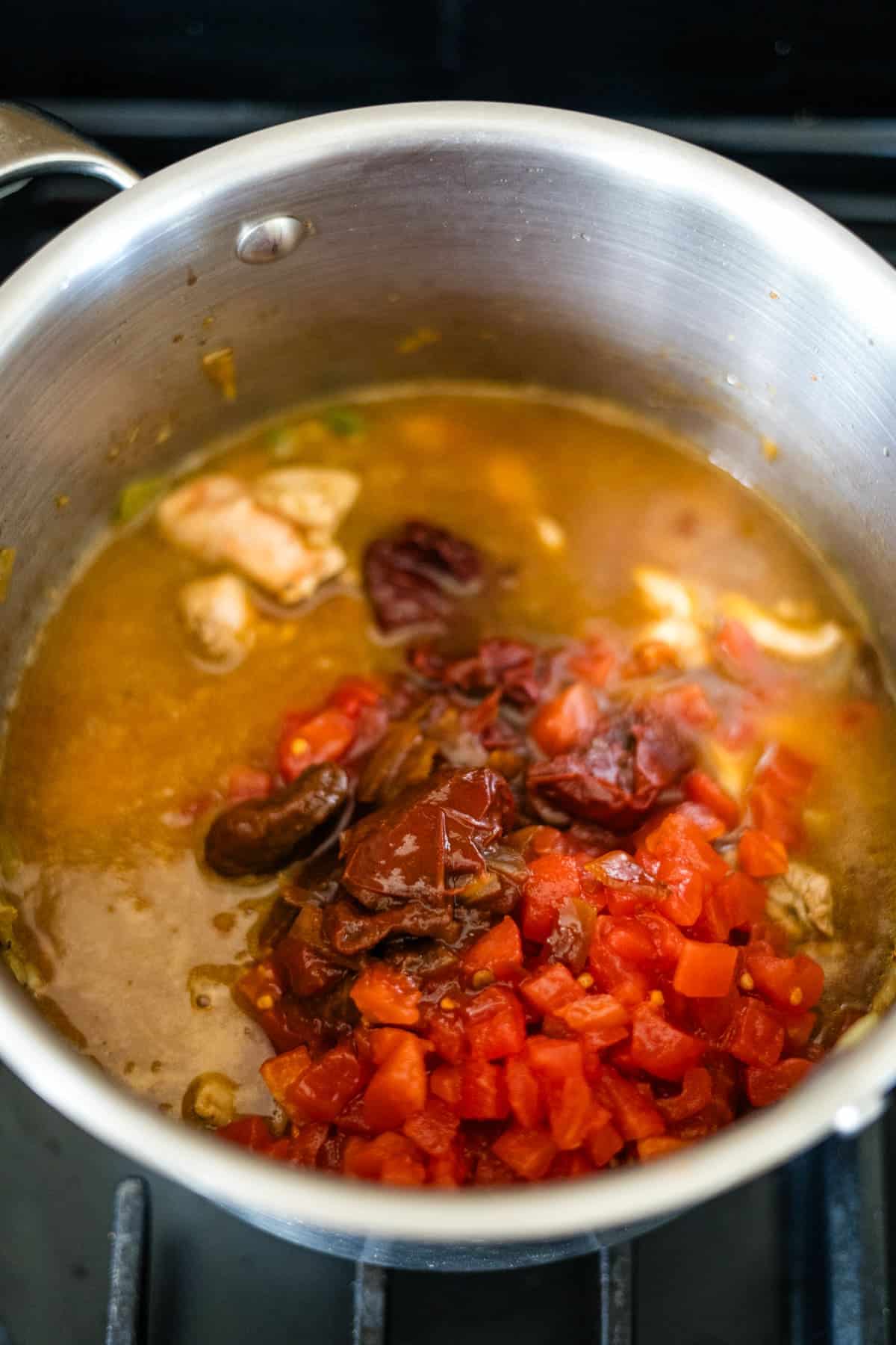 A pot on a stove with chopped tomatoes, dried peppers, and chunks of meat mingling in a simmering chicken chipotle soup.