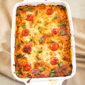 Cheesy baked casserole with cherry tomatoes and herbs in a white dish on a textured cloth surface.