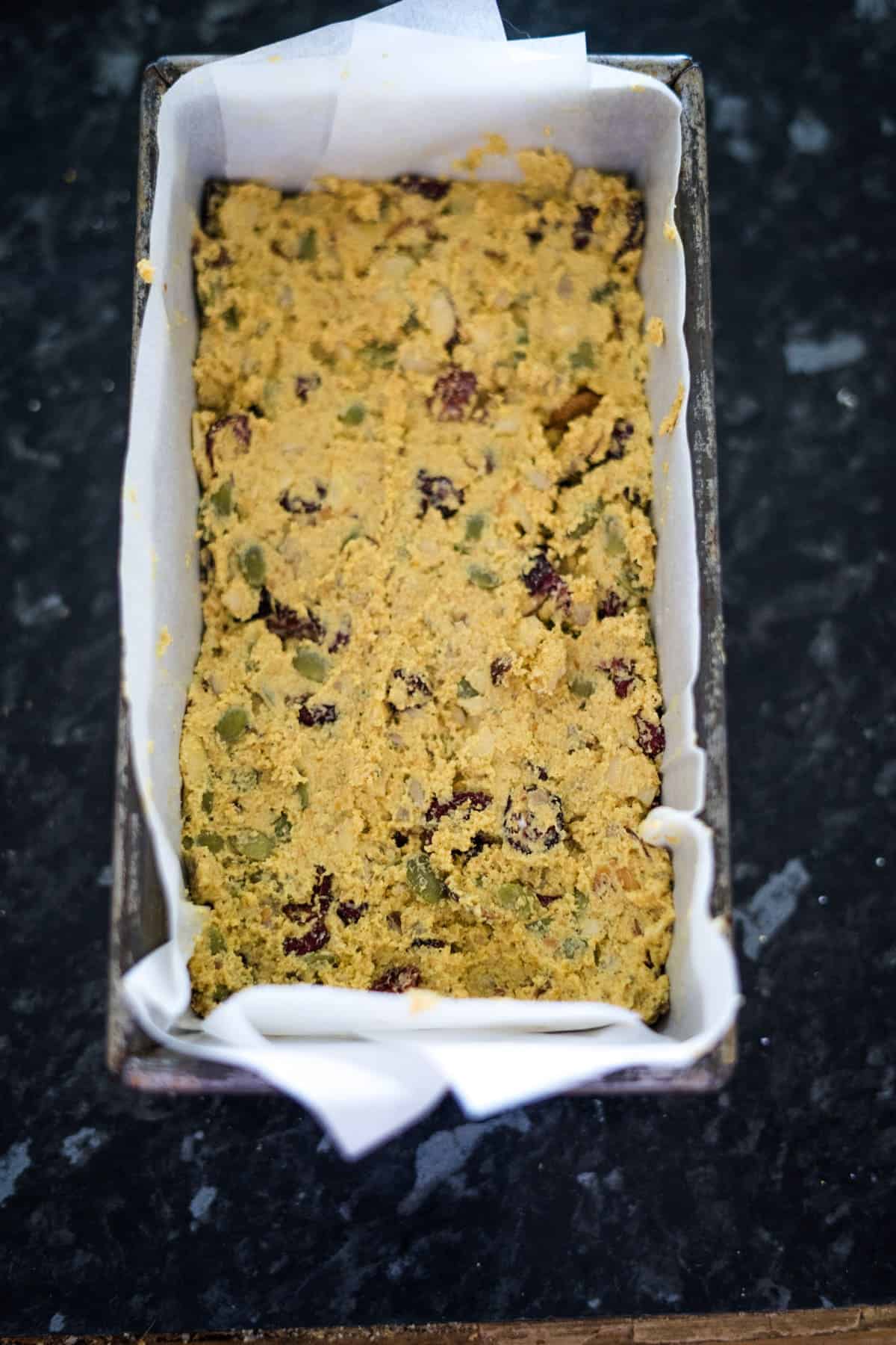 A loaf pan with unbaked cornbread mixture, lined with parchment paper, containing visible seeds and dried cranberries.