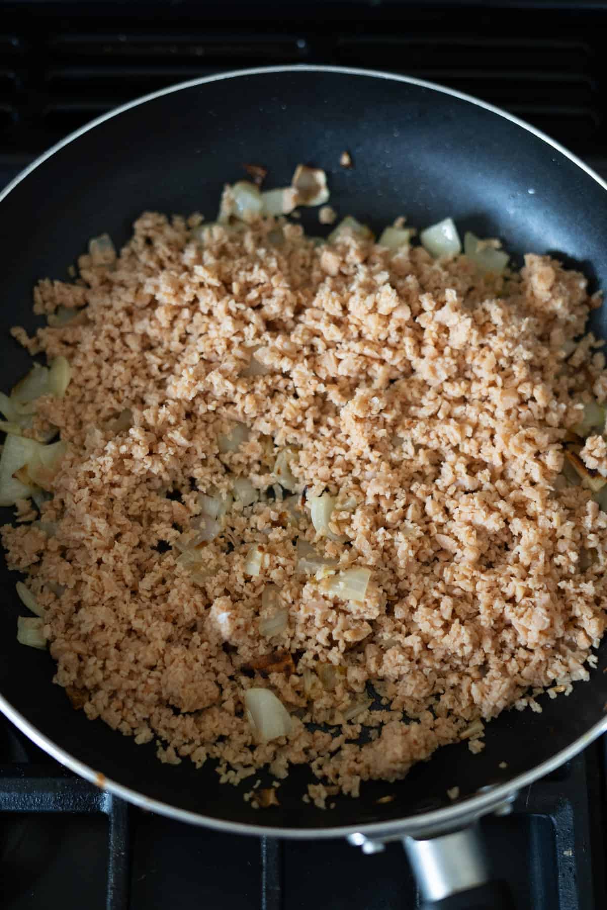 A frying pan on a stove contains cooked ground meat and chopped onions.