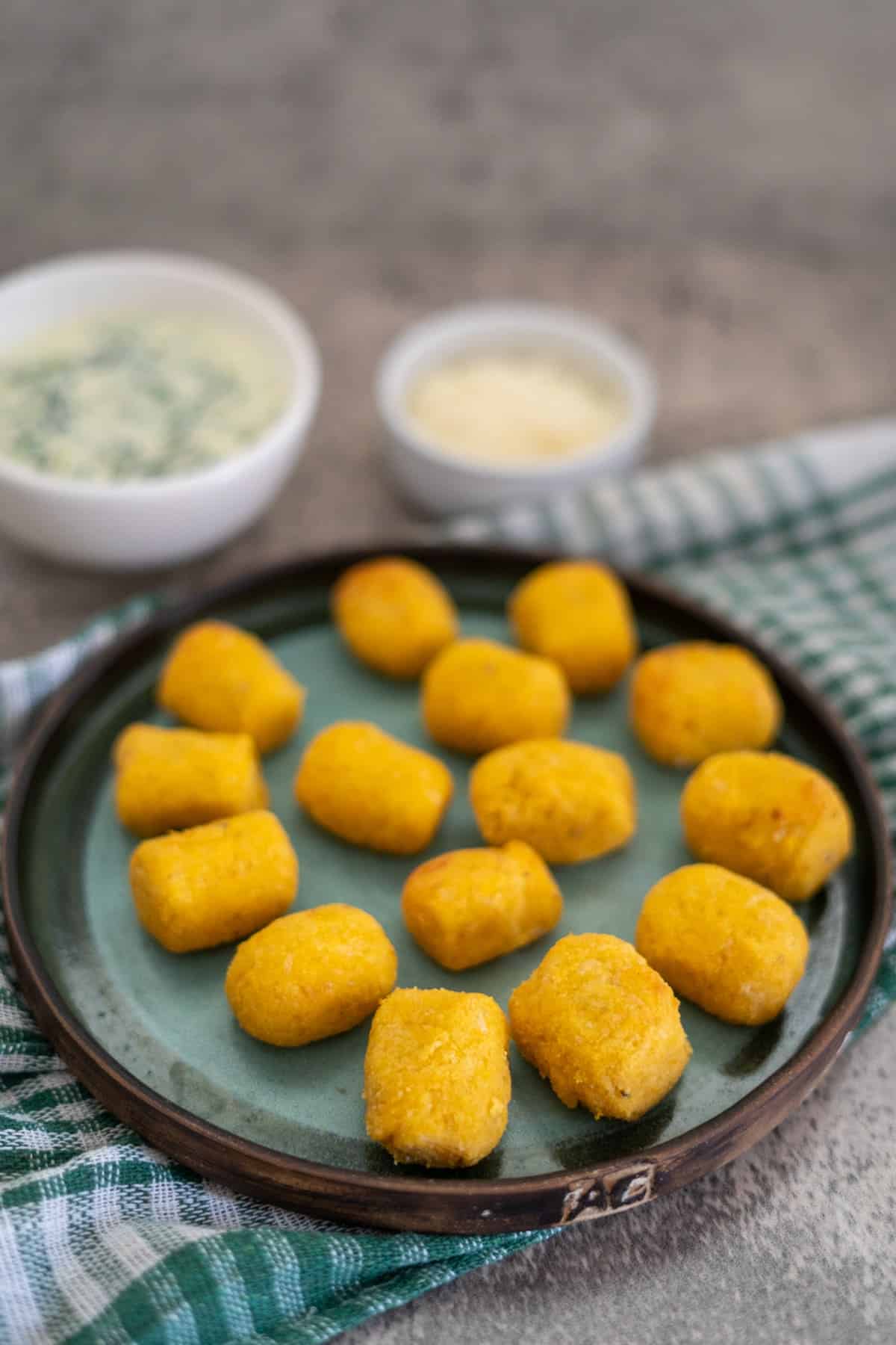 A plate of golden fried croquettes on a green dish rests atop a checkered cloth, with two small bowls of dipping sauces as an enticing accompaniment. Nearby, a serving of gluten-free pumpkin gnocchi awaits to complete the delightful spread.