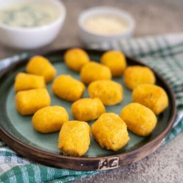 A plate of golden fried croquettes arranged neatly, with dipping sauces in small bowls in the background on a checkered cloth.