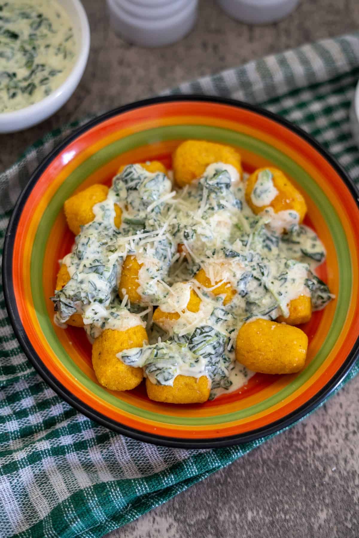 A colorful plate of gluten-free pumpkin gnocchi topped with a creamy spinach sauce, garnished with grated cheese. A green-striped cloth is underneath the plate.