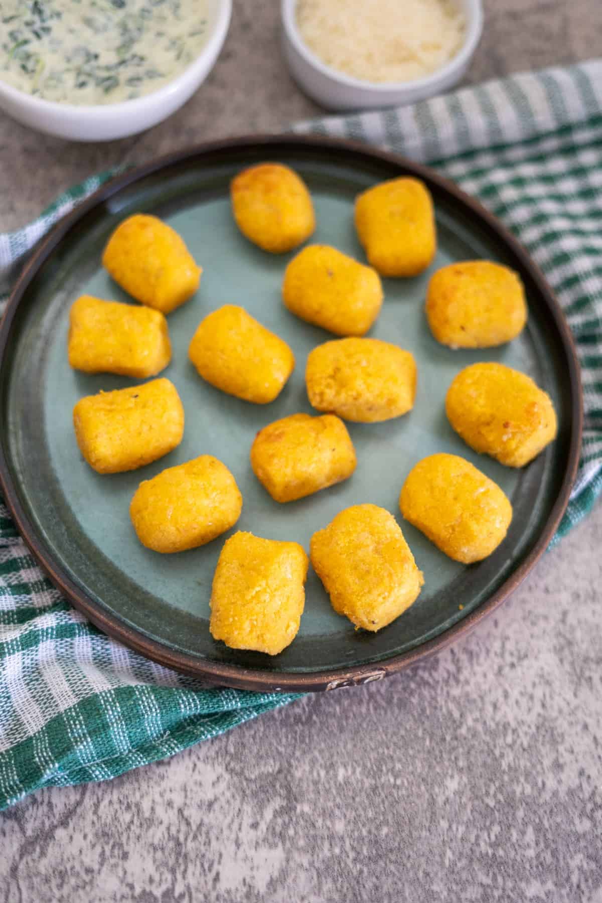 A round plate with golden-brown tater tots on a green and gray striped napkin. Bowls of dip and grated cheese are in the background on a textured surface.