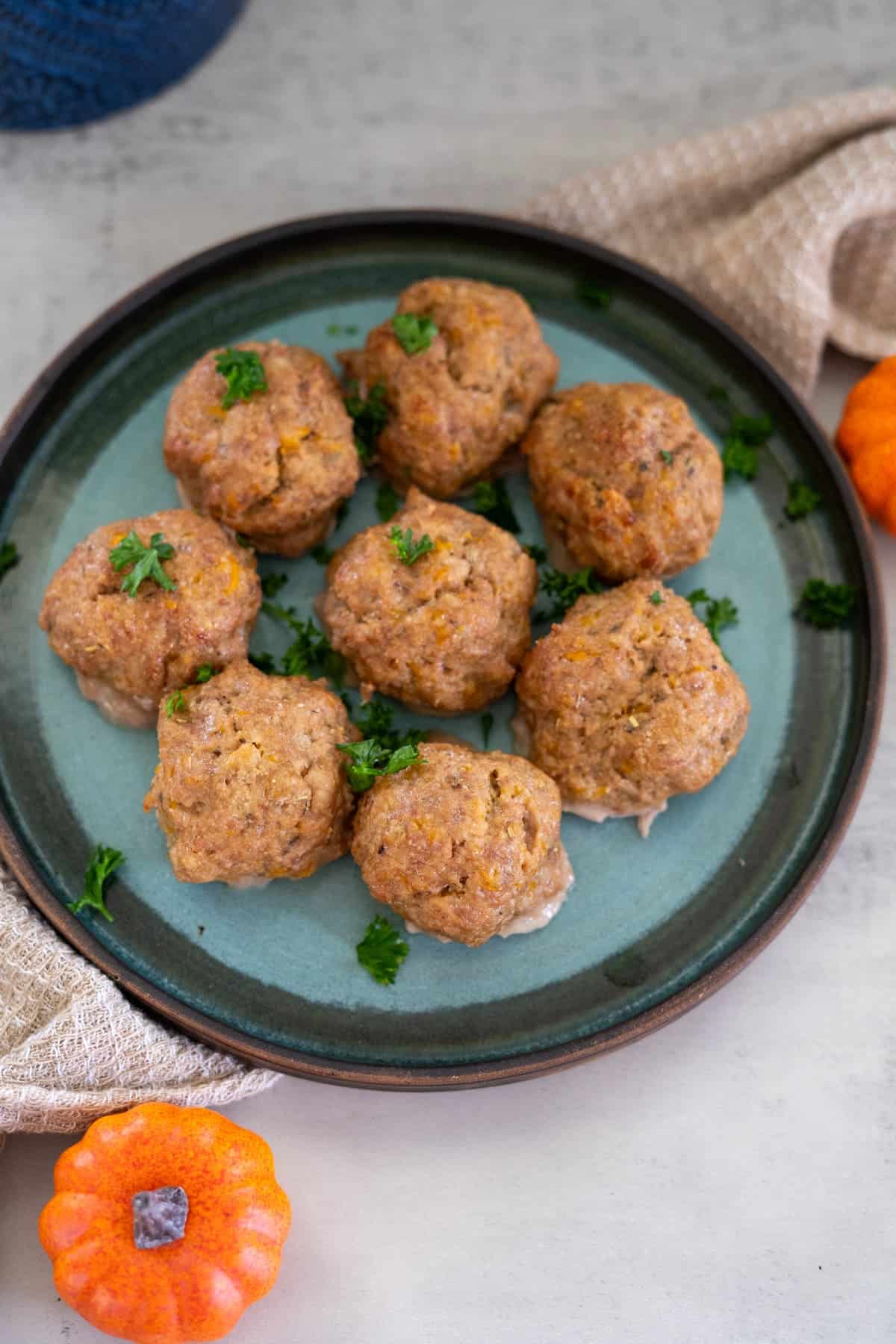 A blue plate with eight baked stuffing balls garnished with parsley, placed on a table next to small decorative pumpkins and a beige cloth.