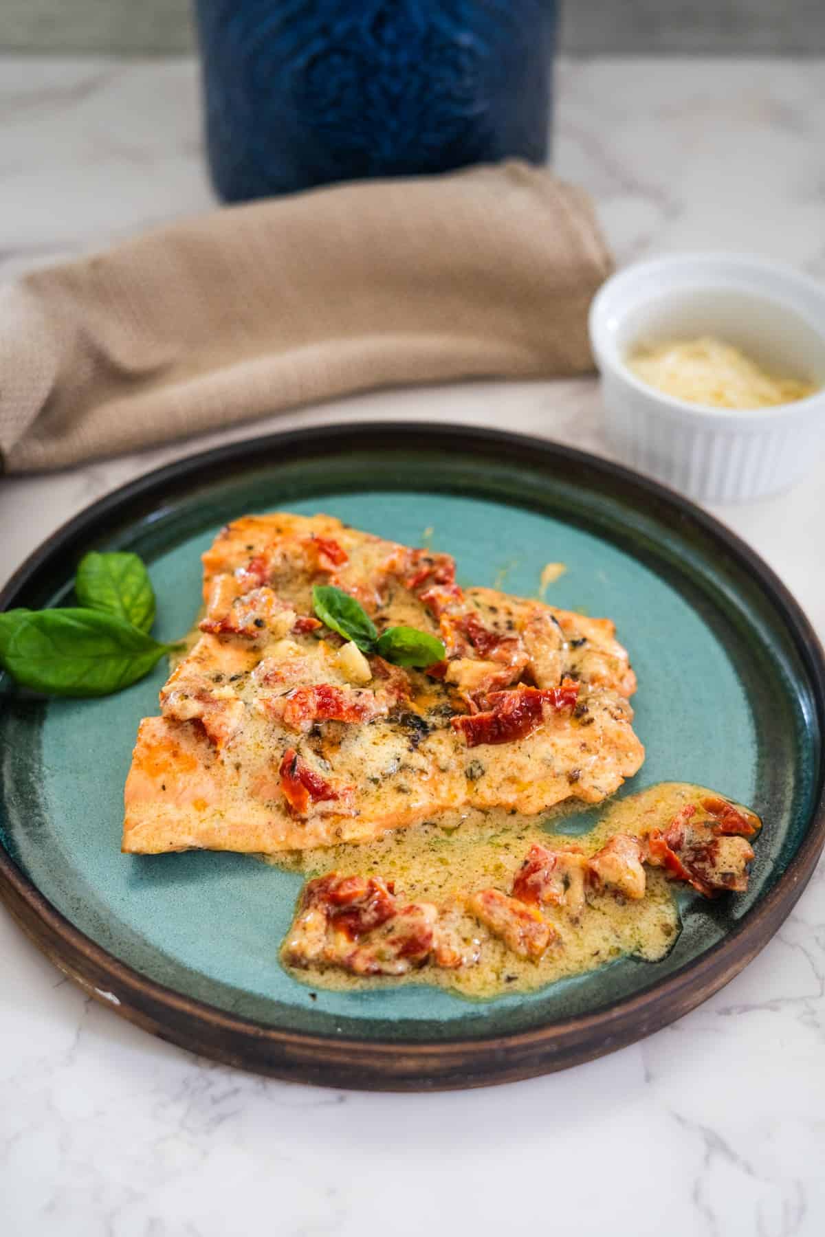 A salmon fillet topped with a creamy sauce and sun-dried tomatoes on a blue-green plate, garnished with basil.