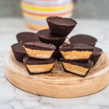 A wooden platter with stacked chocolate peanut butter cups on a marble surface. A striped cup sits in the background.