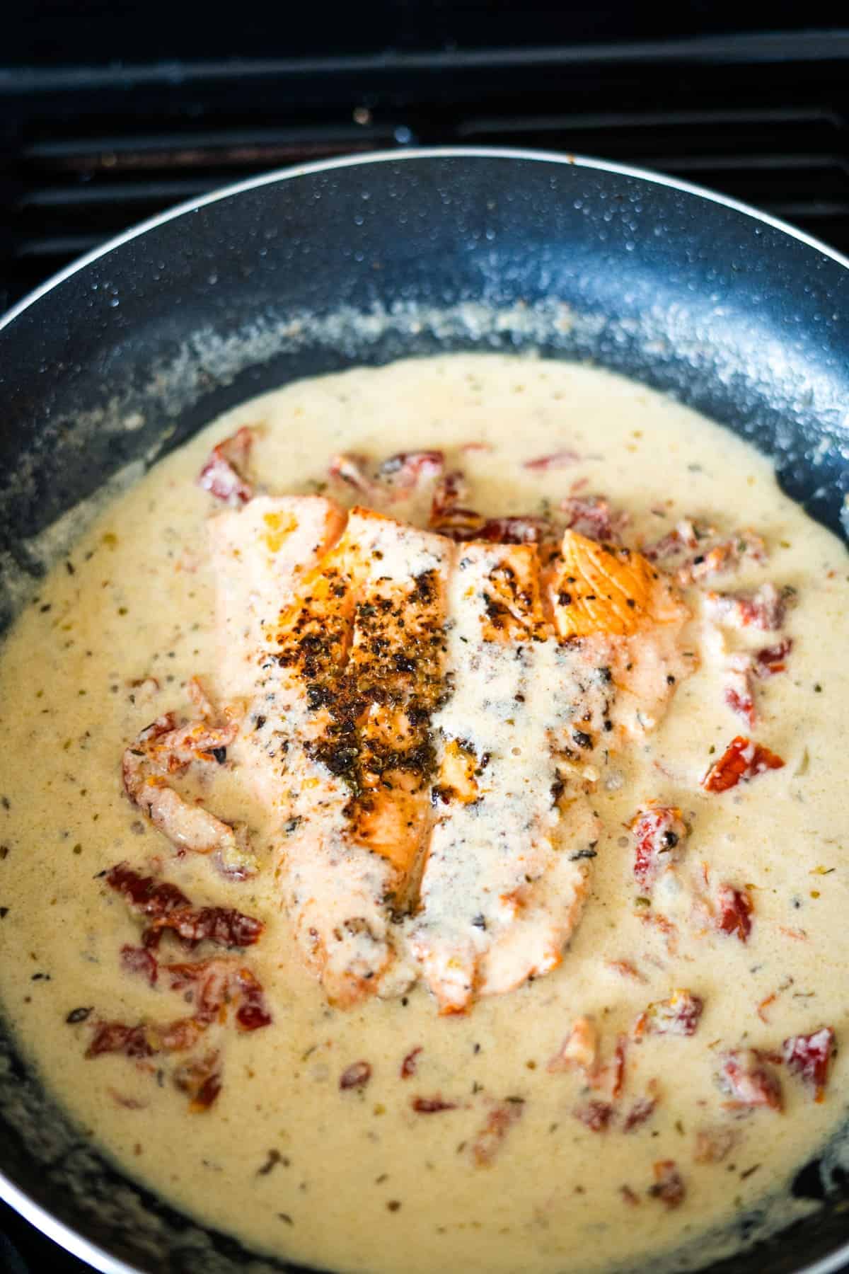 Pan-seared salmon in a creamy sauce with sun-dried tomatoes, cooking in a black skillet on a stove.