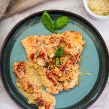 Creamy tomato basil chicken on a blue plate, garnished with fresh basil leaves. A small bowl of grated cheese is nearby.