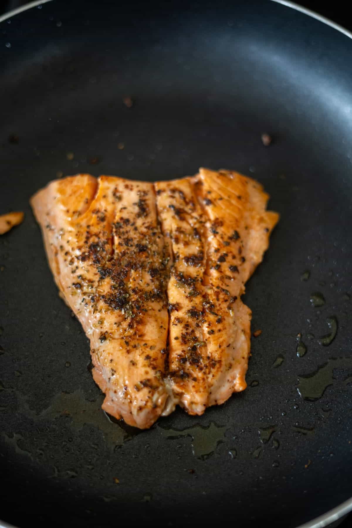 A piece of seasoned salmon is being cooked in a black skillet.