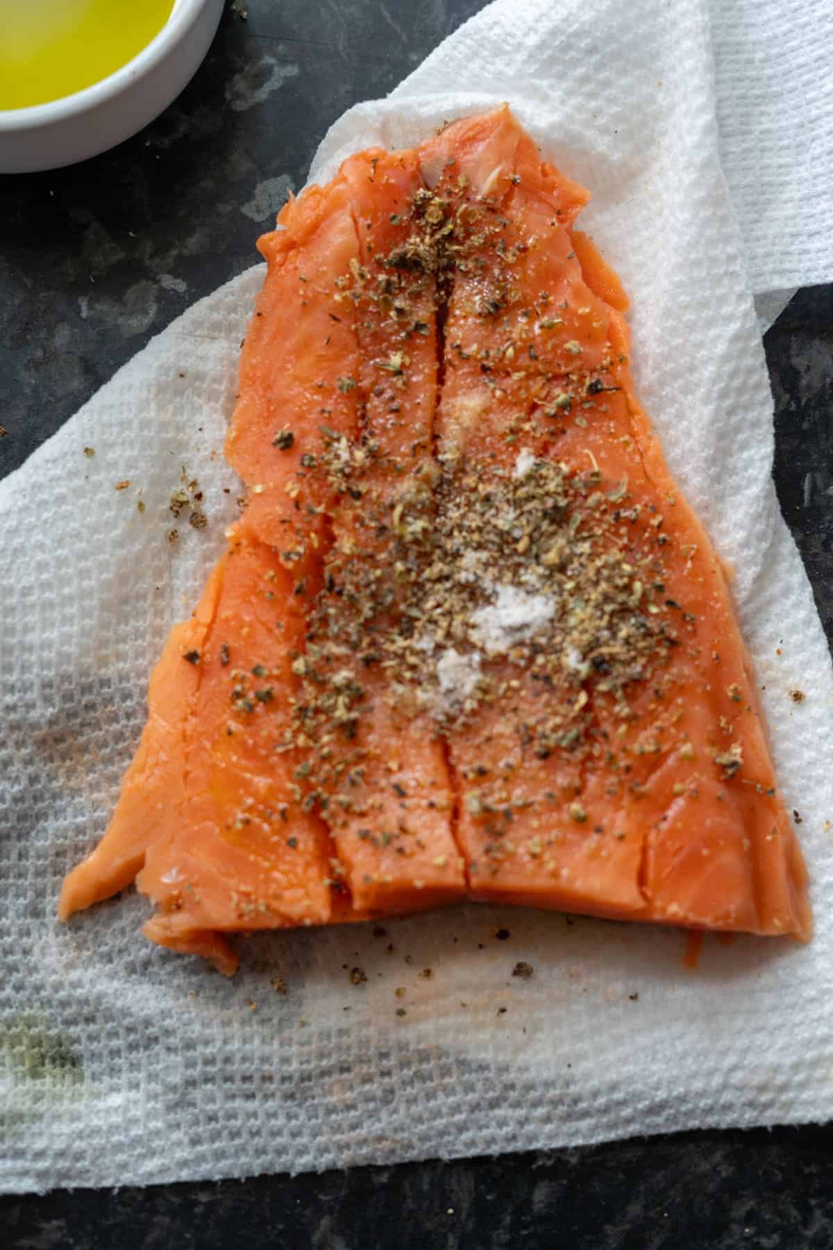 Raw salmon filet seasoned with spices and salt, placed on a paper towel. A small white bowl with a yellow liquid is partially visible in the background.