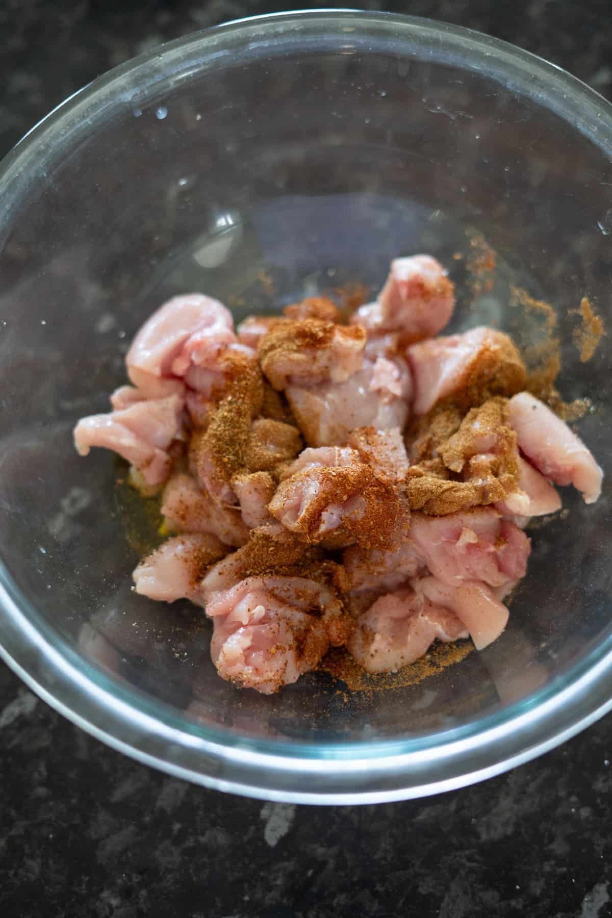 Raw chicken pieces with spices marinate in a clear glass bowl on a dark countertop, perfect for crafting a zesty chicken chipotle soup.