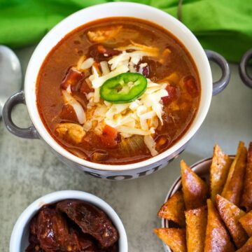 A bowl of chili with shredded cheese and a sliced jalapeño on top, alongside a dish of sliced bread and a small bowl of chipotle peppers.