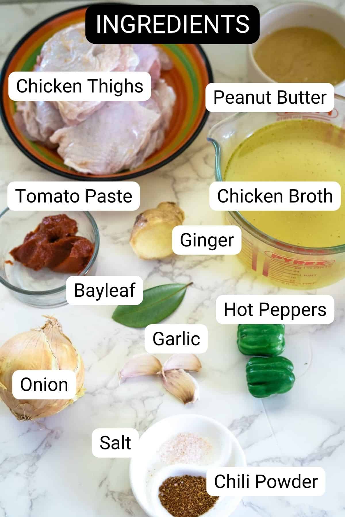 Image of ingredients on a marble surface, including chicken thighs, peanut butter, tomato paste, chicken broth, ginger, hot peppers, onion, garlic, bay leaf, salt, and chili powder.