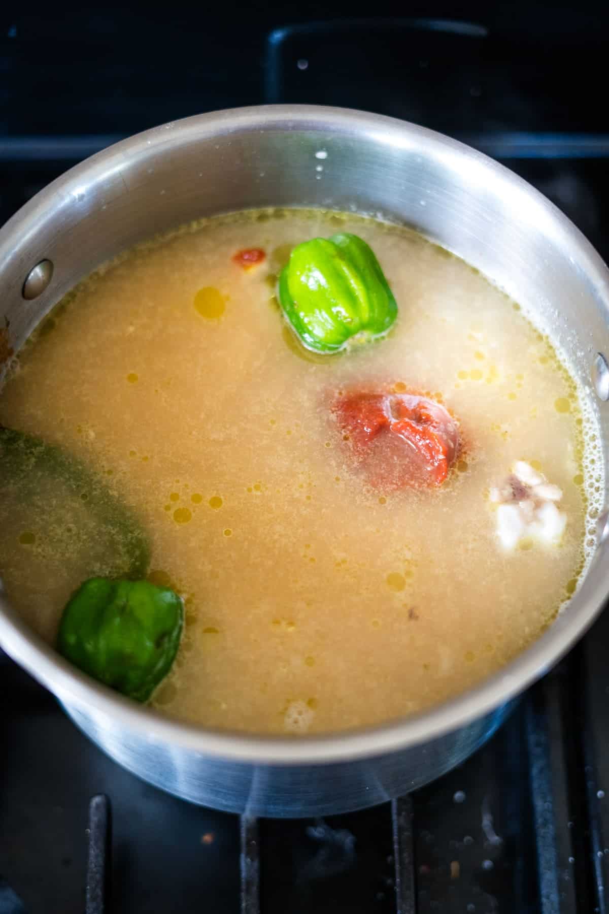 A pot of soup broth with whole green peppers, a tomato, and other ingredients simmering on a stove.