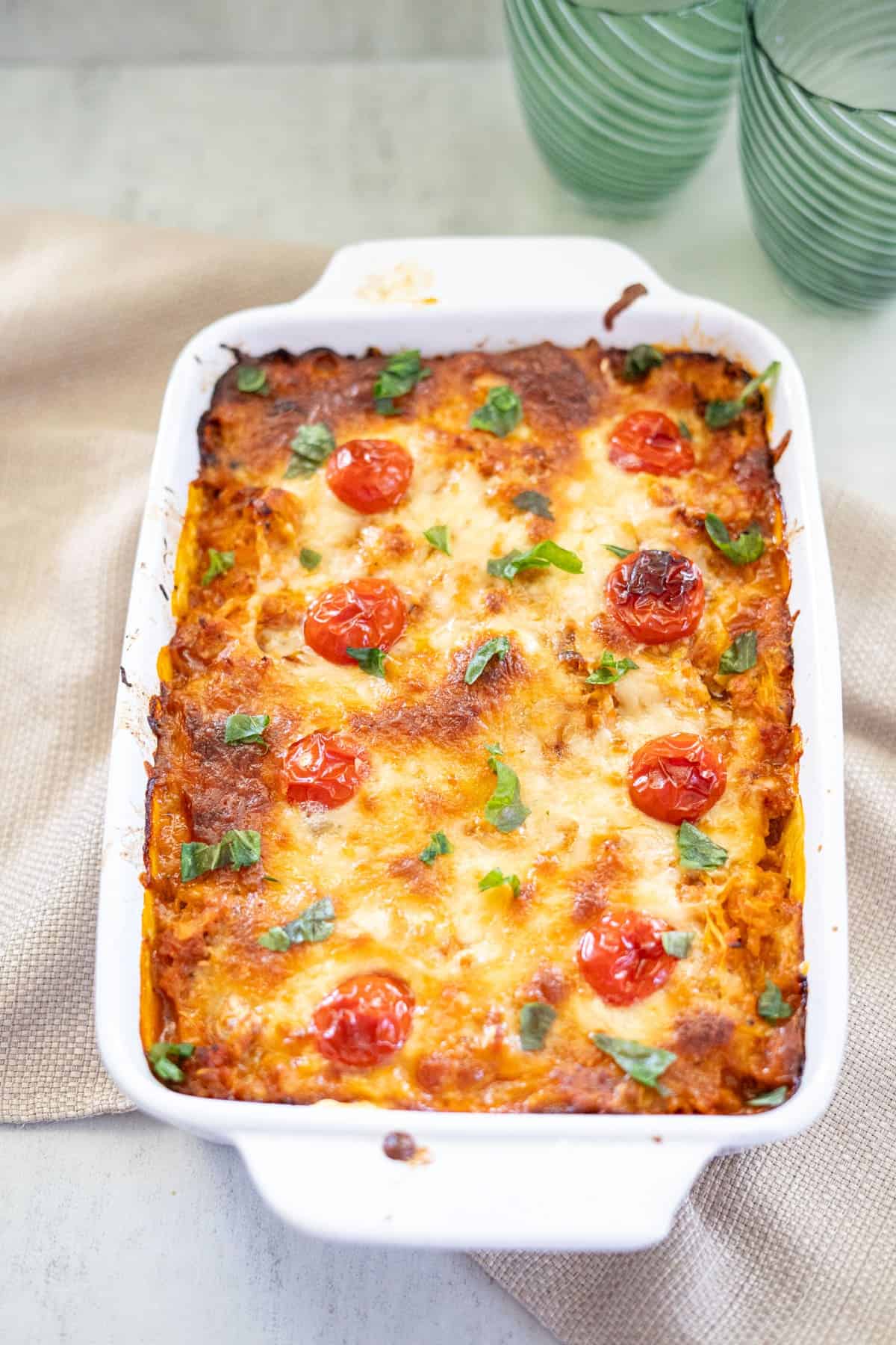 Baked casserole in a white dish with a golden-brown top, garnished with cherry tomatoes and herbs. Two green glasses are in the background.