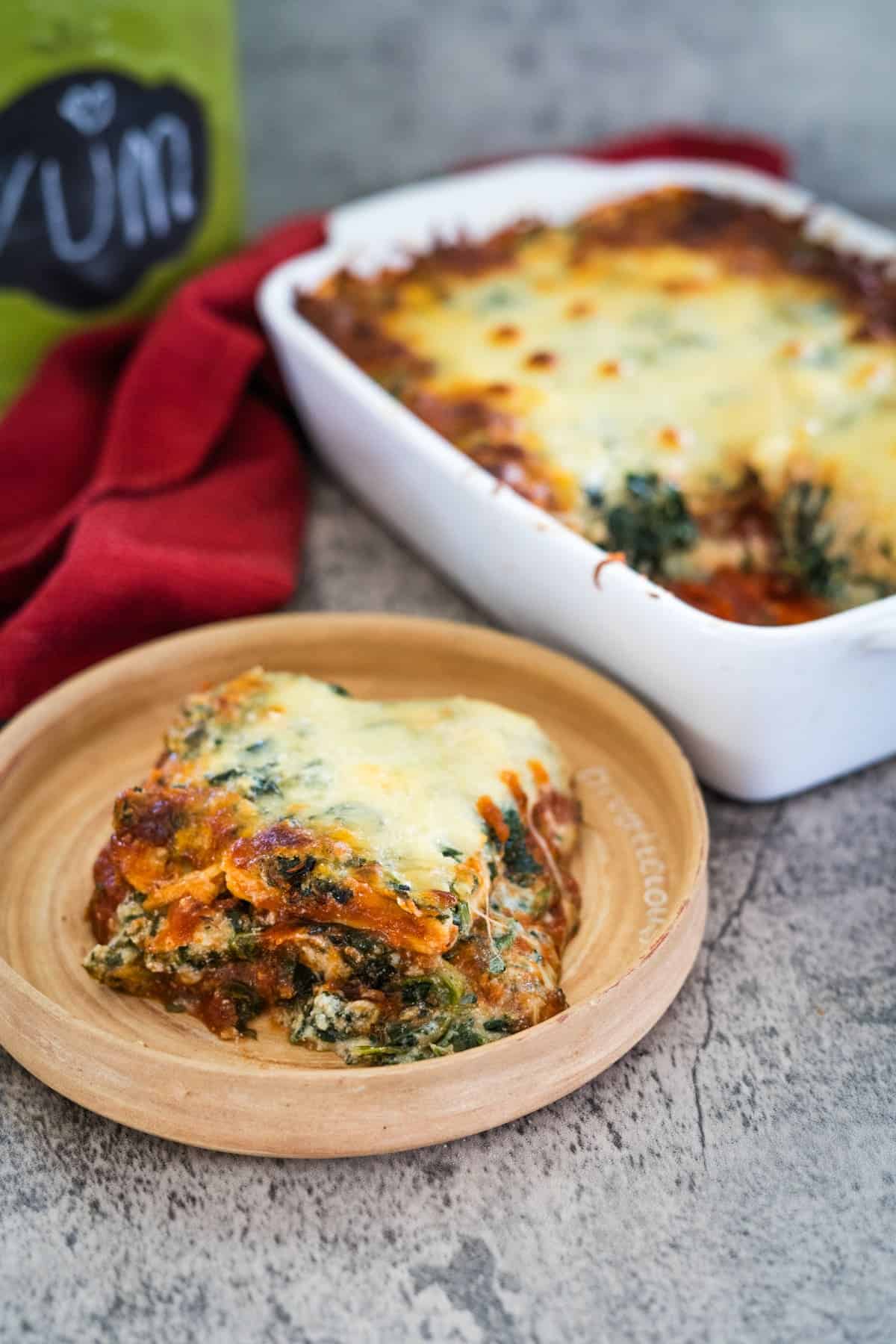 A slice of keto spinach lasagna rests on a wooden plate, with a baking dish of the deliciously healthy lasagna in the background, nestled next to a red cloth and a sign reading "Yum.