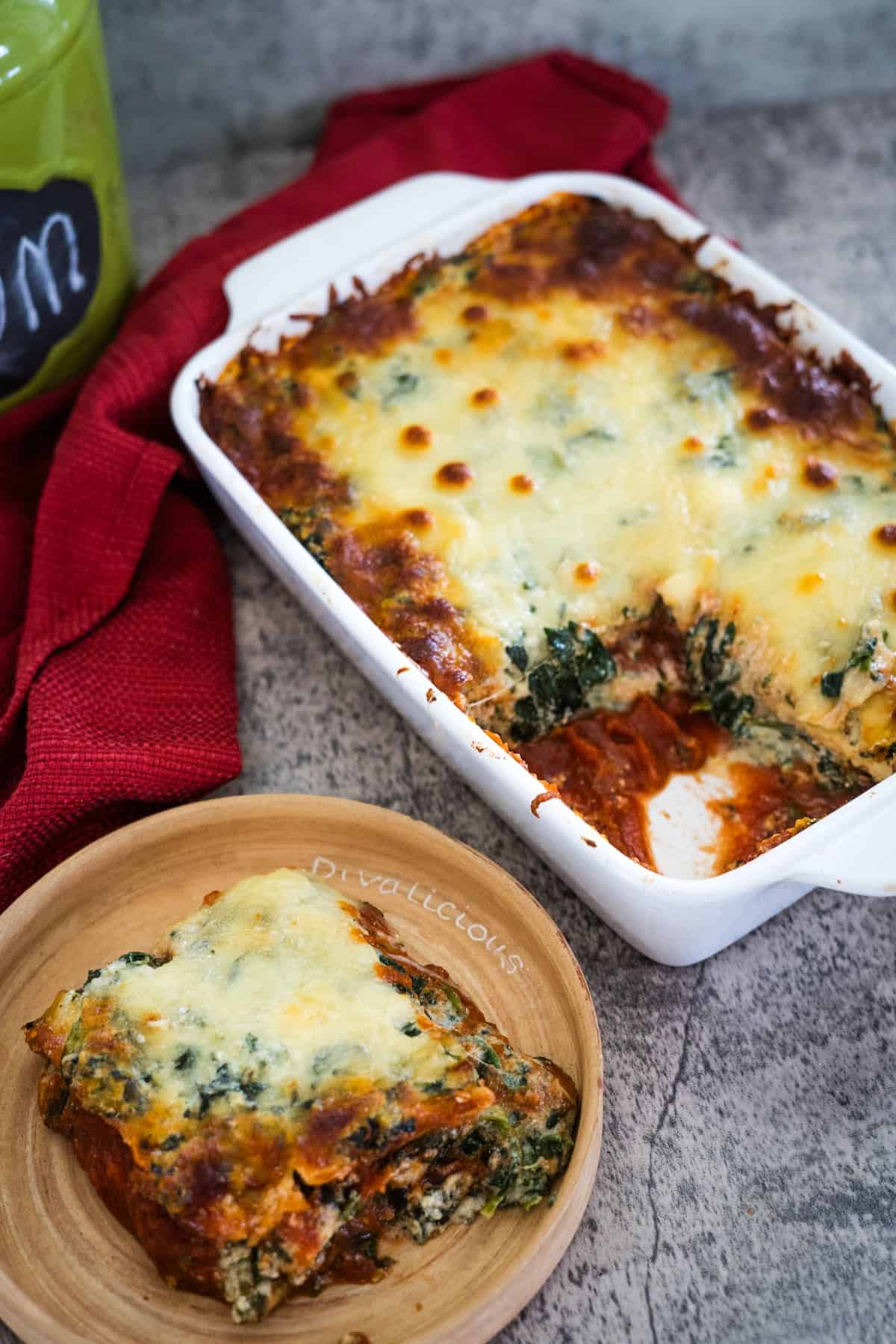 Baked casserole dish with a serving of cheesy spinach lasagna on a plate, next to a red cloth.