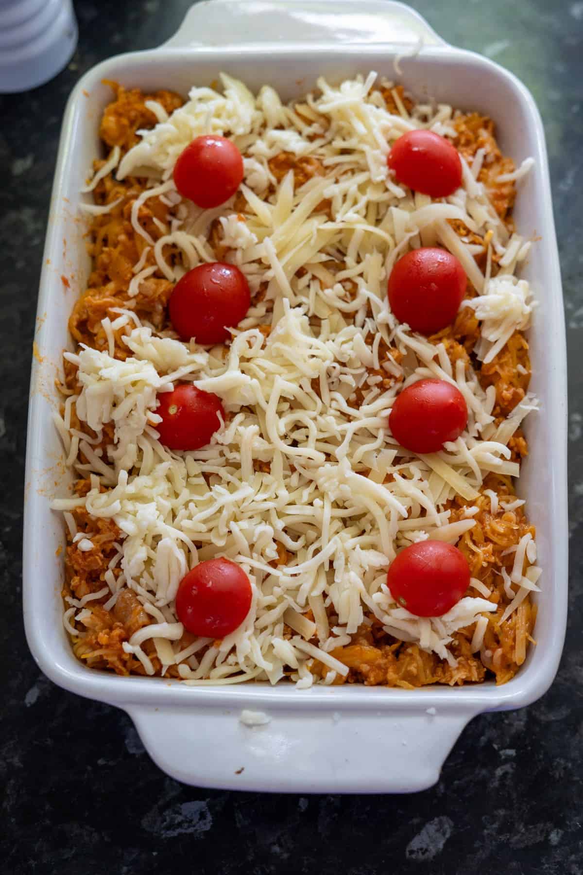 Baking dish with shredded cheese, cherry tomatoes, and shredded sweet potatoes on a dark countertop.