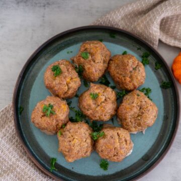 A plate with eight round meatballs garnished with parsley, set on a textured beige cloth.