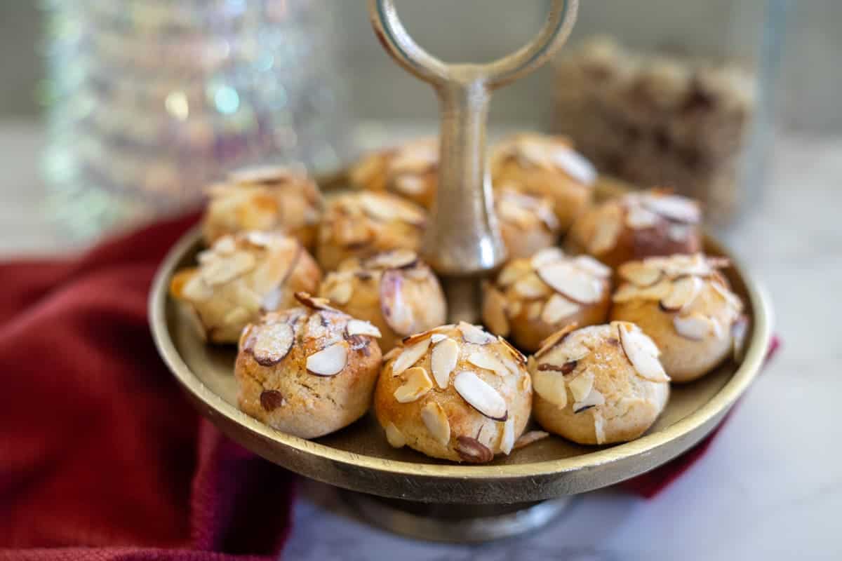 A plate of almond cookies, crowned with almond slices, rests gracefully on a pedestal stand beside a red cloth.