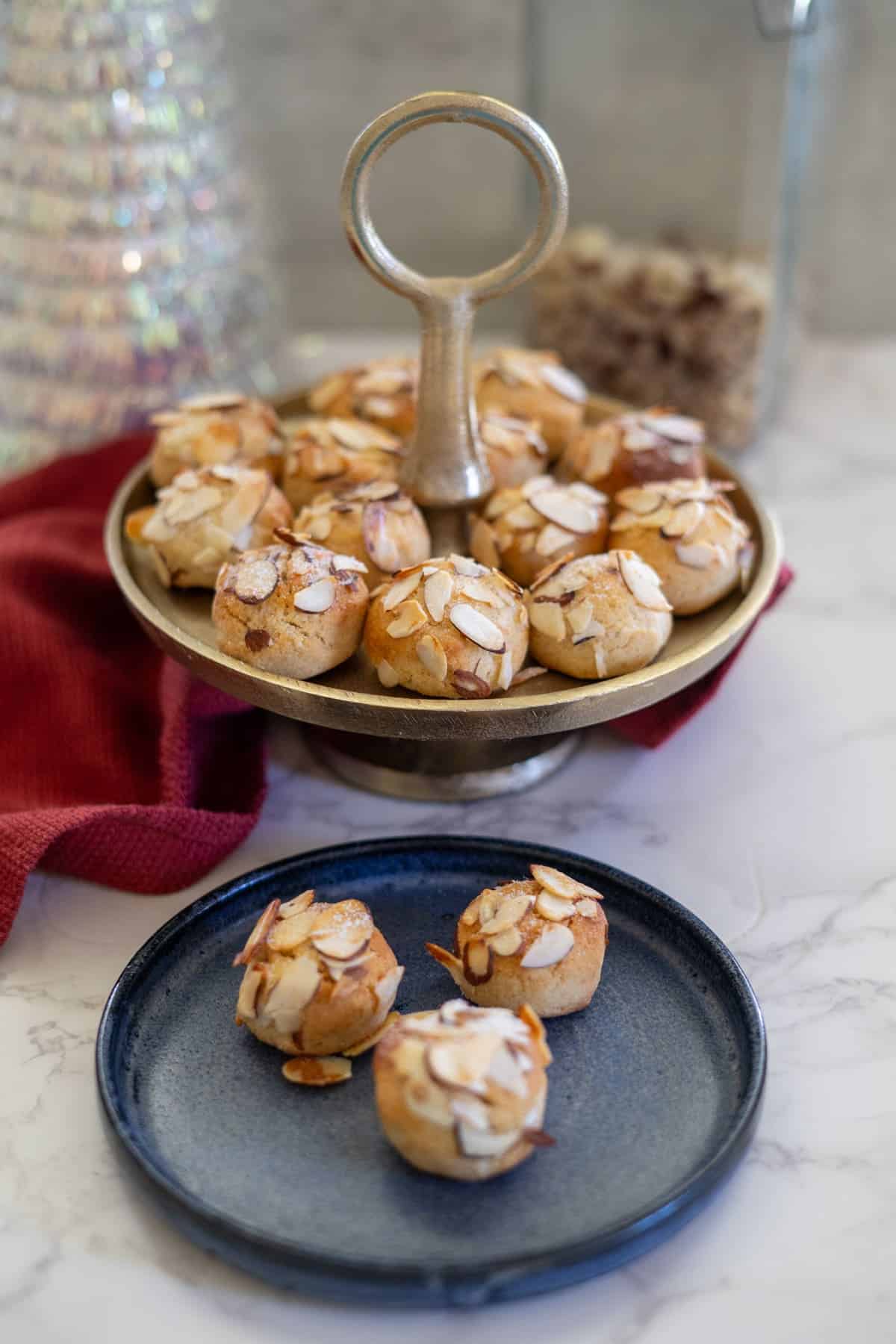 A charming display of almond cookies graces the two-tiered stand, with three delicately perched on a blue plate. A rich red cloth and a glass jar provide an elegant backdrop to this delightful scene.