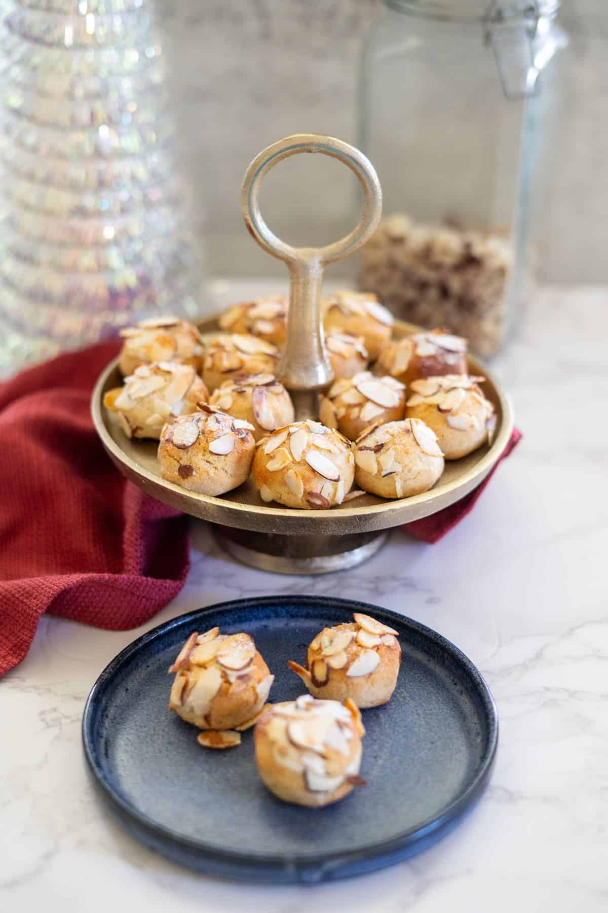 Small almond pastries, or almond cookies, are displayed on a tiered tray, with more on a blue plate. A red cloth and a glass jar provide a charming backdrop on the marble surface.