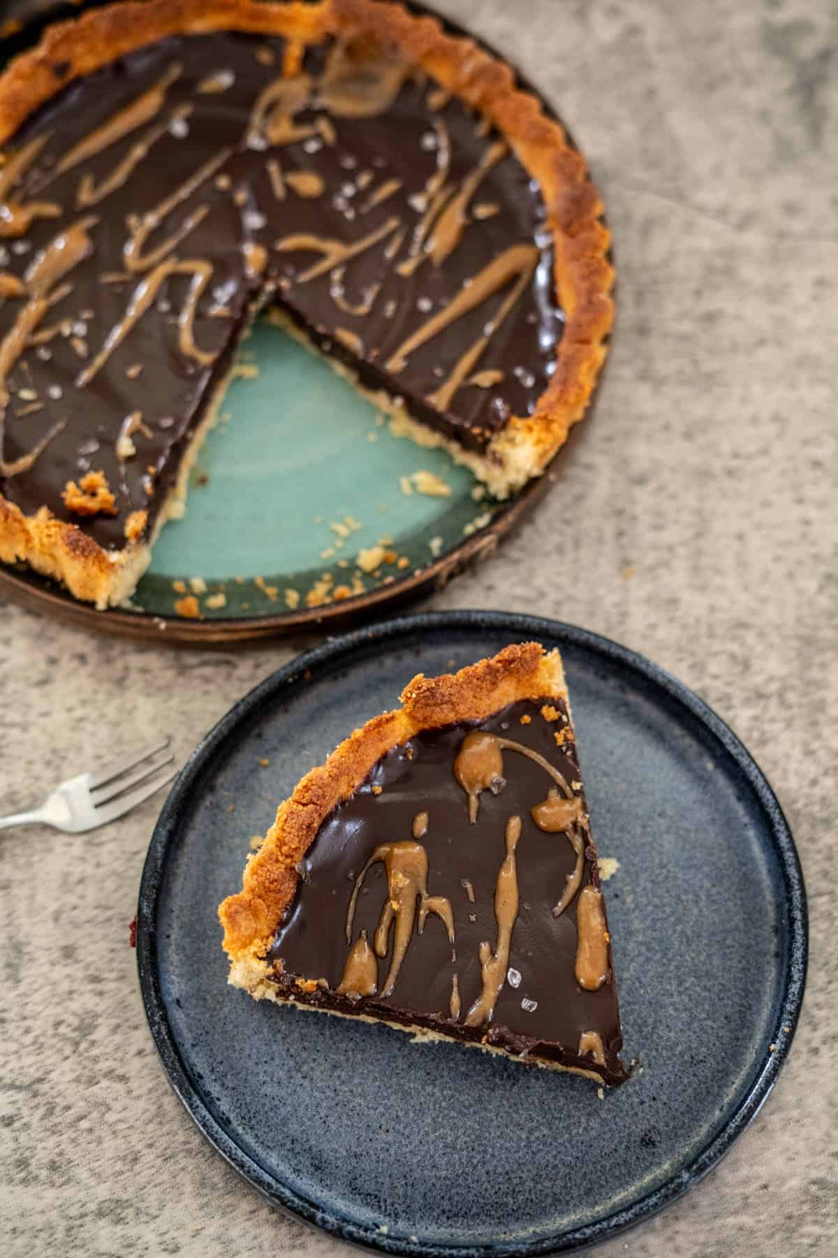 Slice of chocolate tart with caramel drizzle on a blue plate, and the remaining tart with missing slice on a green platter. Fork nearby on a gray surface.