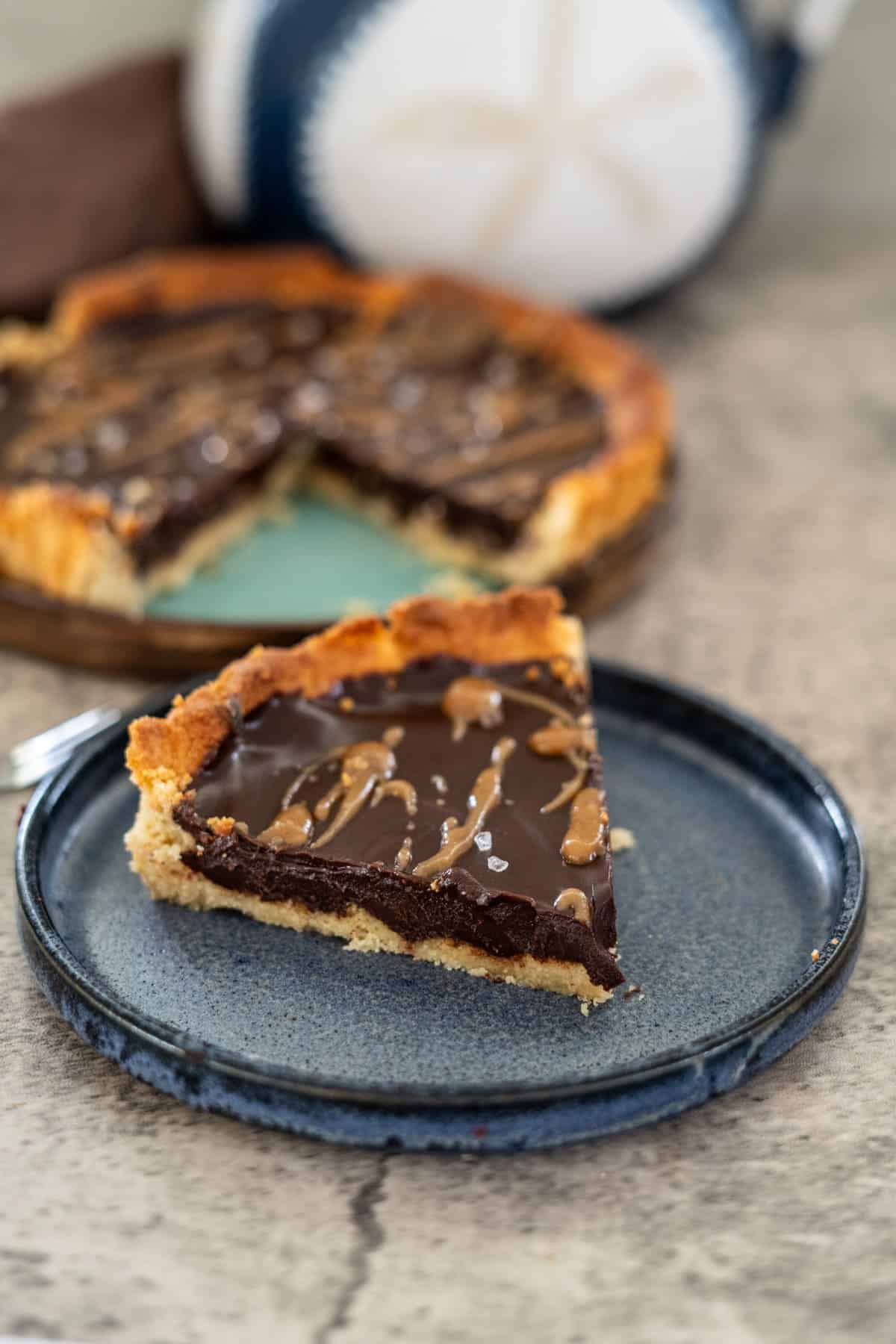 A slice of keto chocolate tart with caramel drizzle on a blue plate. The rest of the tart is in the background on a green plate, showcasing its decadent allure.