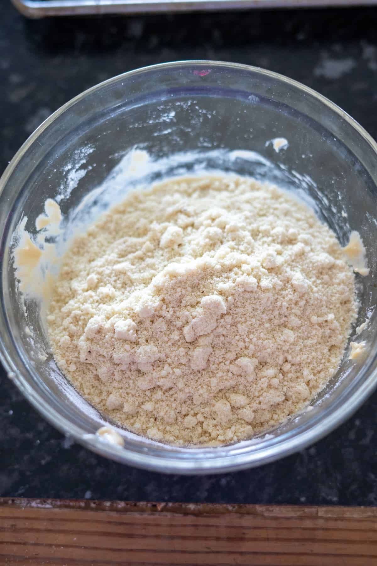 A glass bowl containing a crumbly, beige dough mixture sits on a dark countertop, reminiscent of the textures in keto Twix recipes.