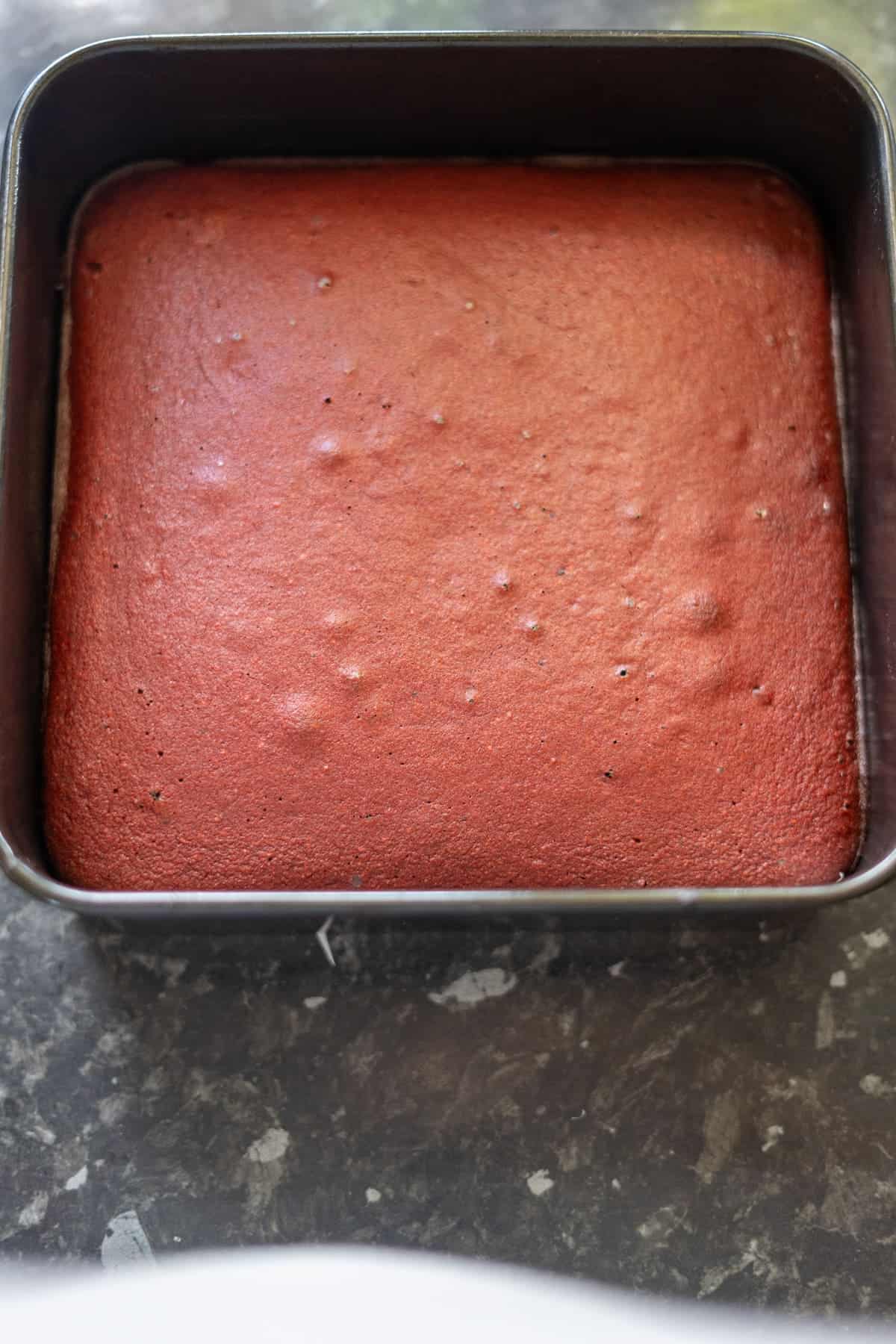 A square red velvet cake in a dark baking pan on a marble countertop.