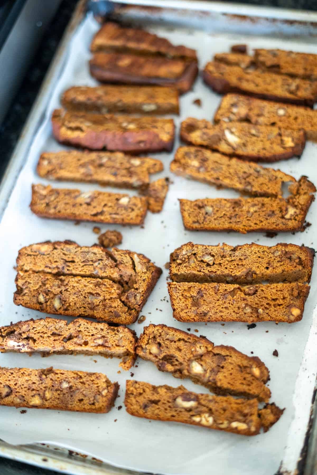 A baking sheet with multiple slices of pumpkin biscotti, placed on parchment paper. Each piece is golden brown with visible chunks of nuts.