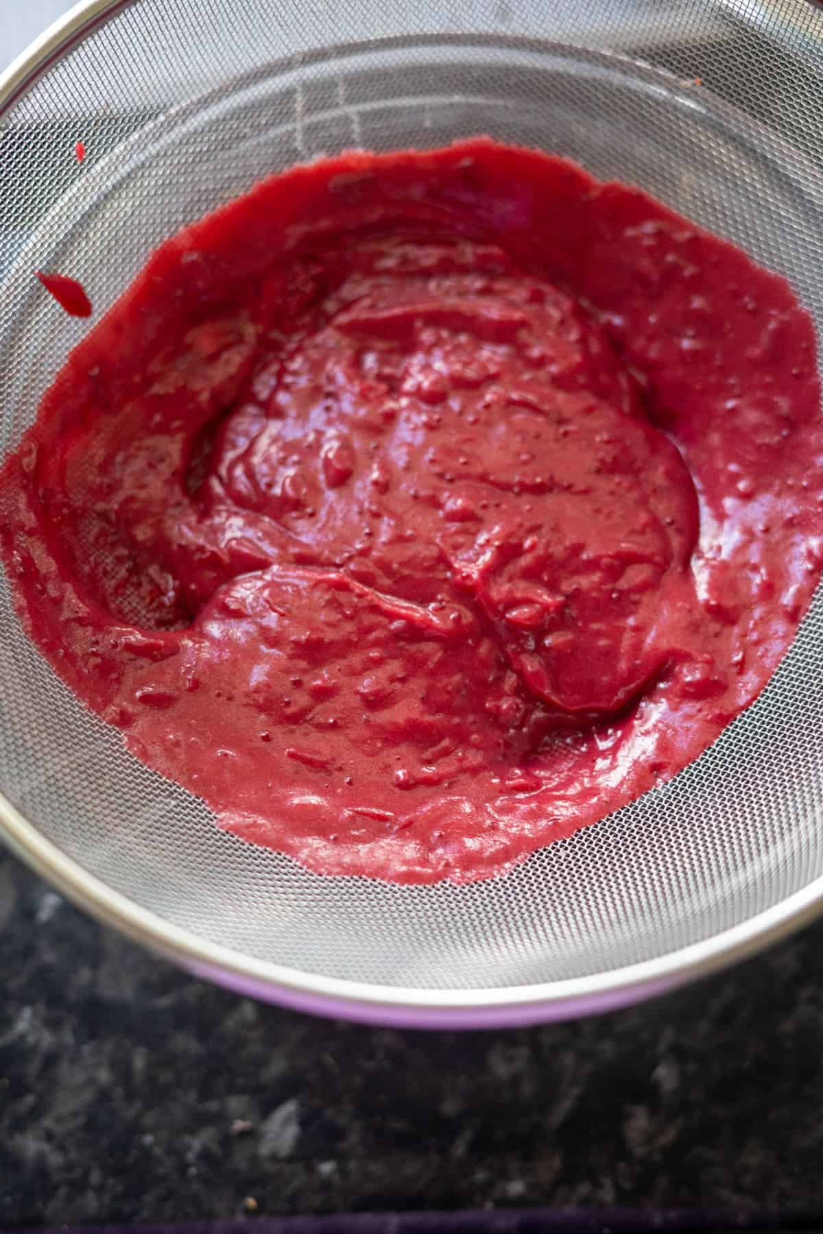 A thick red mixture is strained through a metal mesh sieve over a black countertop.