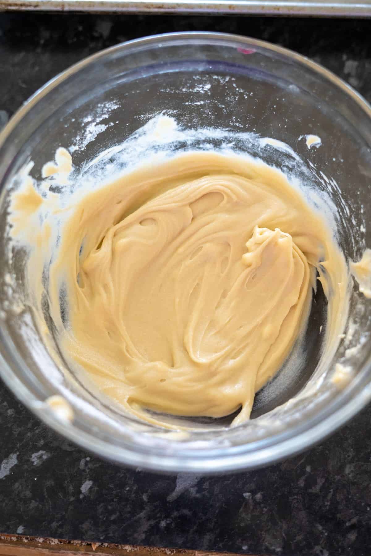 A glass bowl containing smooth, light-colored batter, perfect for crafting a keto Twix treat, rests on a dark countertop.