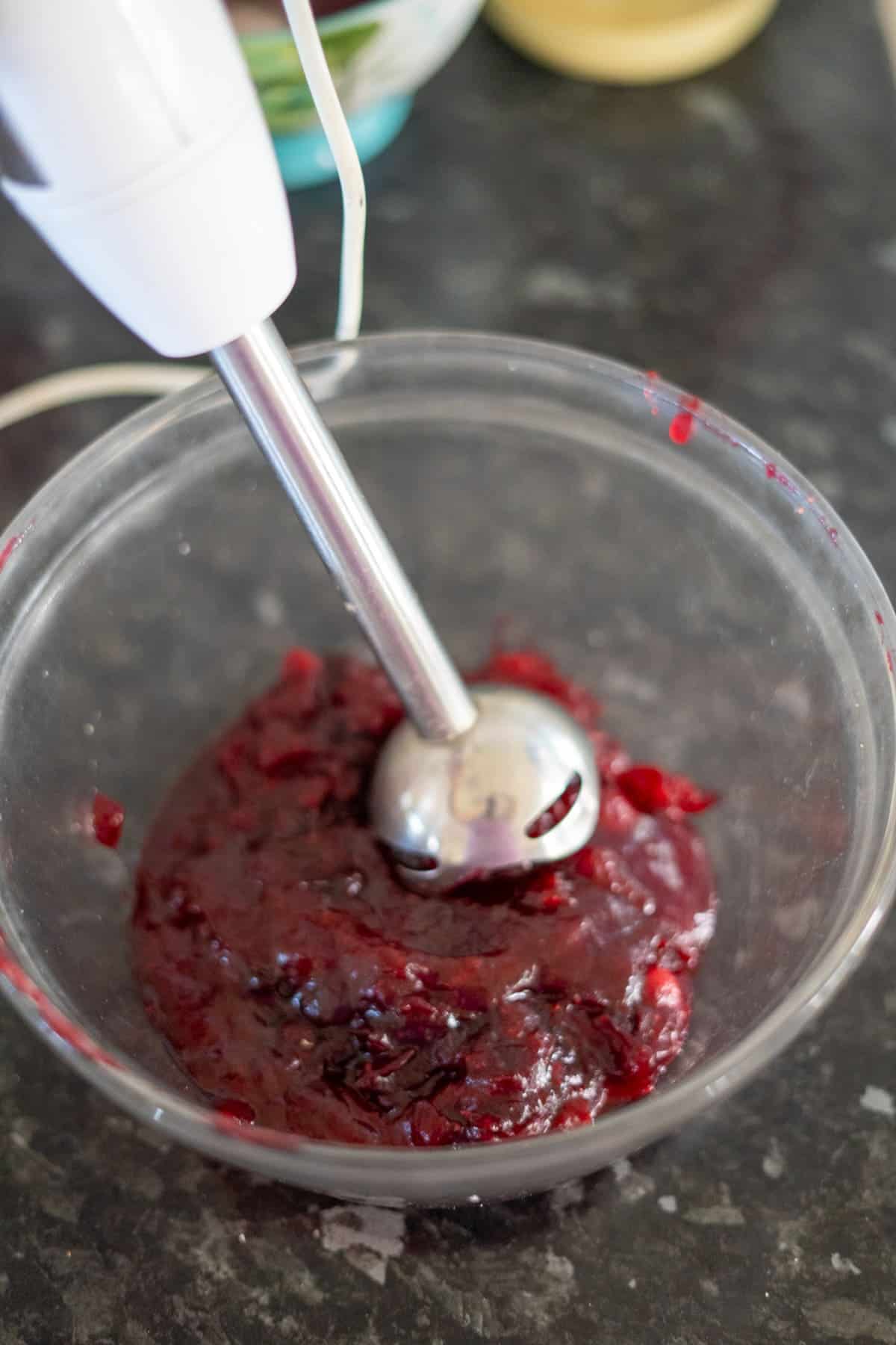 An immersion blender is mixing a dark red fruit mixture in a clear bowl on a kitchen counter.