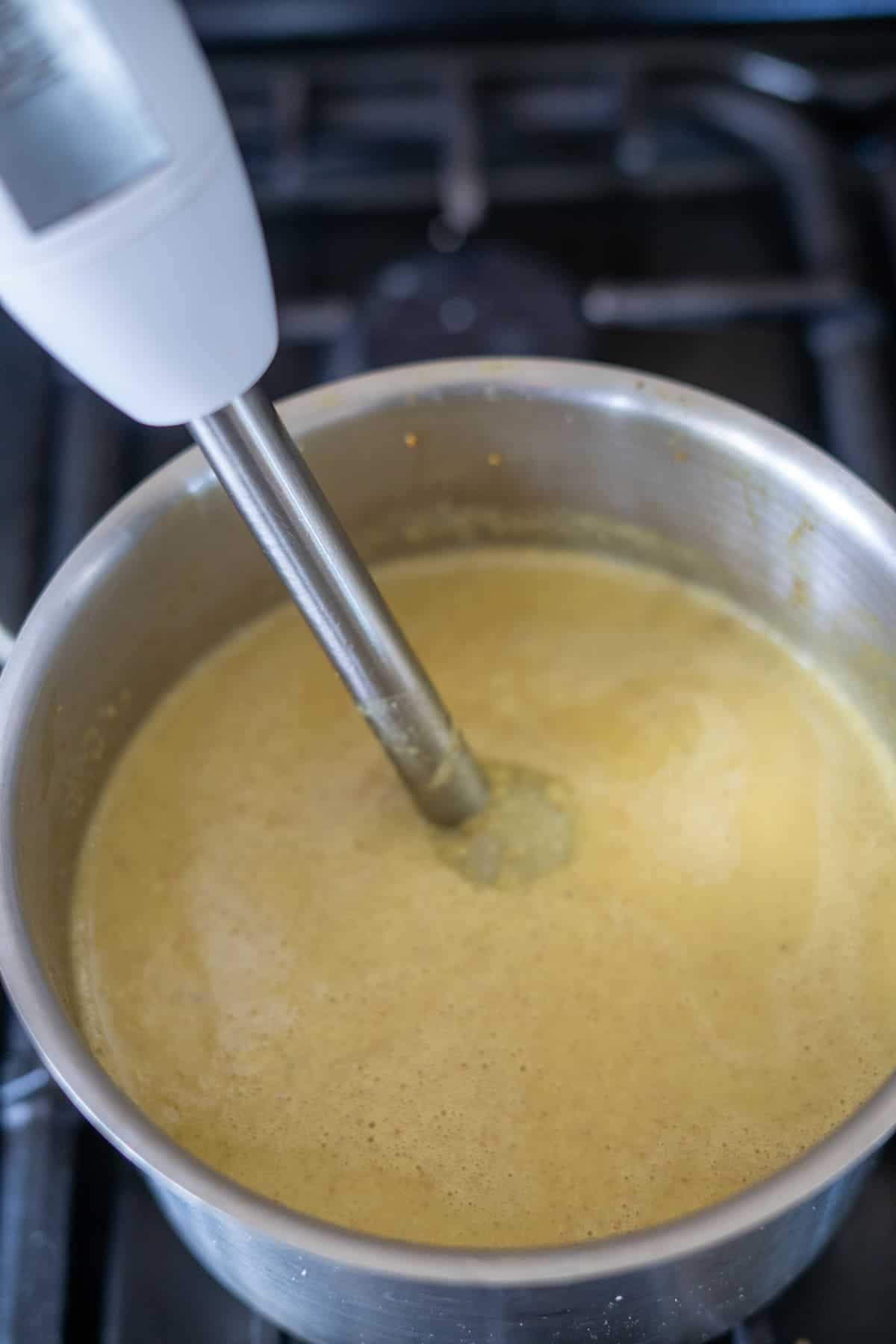 An immersion blender is blending a mixture in a stainless steel pot on a stove.